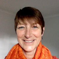 Close up portrait of Cathie smiling. Wearing an orange scarf – orange is the colour of the second (sacral) chakra representing water and being in flow with life.