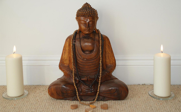 Wooden seated Buddha between 2 pillar candles with 3 small wooden hearts. Represents a return to stillness and peace in the body, breath and mind through meditation and by holding attention in the heart space. 